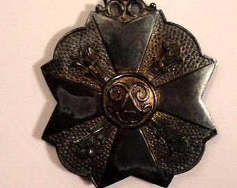 Könnte beinhalten: Eine silberne und goldene Medaille mit einem Sternendesign und einem rot-schwarz gestreiften Band. Die Medaille hat ein zentrales Design eines stilisierten Knotens.