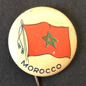 May include: A vintage button with a red flag featuring a green five-pointed star. The flag is on a white background with the word "MOROCCO" printed in blue below the flag.
