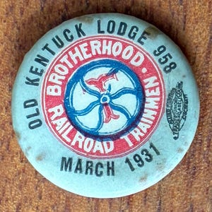 May include: A vintage round pin with a light blue background. The pin features the text "Old Kentucky Lodge Brotherhood of Railroad Trainmen" in a circular pattern. The center has a red and blue design with the text "March 1931".