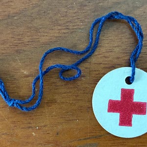 Puede incluir: Una etiqueta redonda, azul claro, con una cruz roja, unida a una cuerda azul. La etiqueta es de papel y tiene un agujero en la parte superior para la cuerda. El fondo es una superficie de madera.