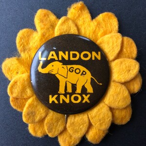 May include: A vintage political campaign button with the text "LANDON KNOX" and a yellow elephant graphic, set against a black background. The button is surrounded by a yellow felt sunflower.
