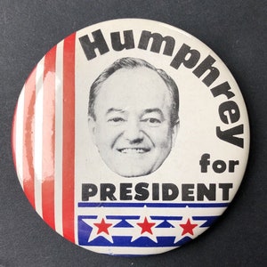 May include: A white button with red, white, and blue stripes around the edge. The button features a black and white photo of Hubert Humphrey, with the text "Humphrey for President" in black lettering.