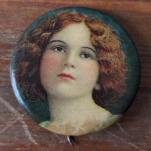 Puede incluir: Botón redondo vintage con un retrato de una mujer con cabello castaño rojizo, mejillas rosadas y labios rojos. El botón tiene un borde verde y un cierre metálico en la parte posterior. La obra de arte recuerda las ilustraciones de principios del siglo XX.