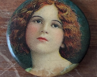 May include: Vintage round button pin featuring a portrait of a woman with auburn hair, rosy cheeks, and red lips. The pin has a green border and a metal clasp on the back. The artwork is reminiscent of early 20th-century illustrations.