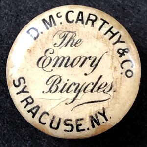 May include: A white button with black text that reads "D.M. McCarthy & Co. The Emory Bicycles Syracuse, NY."