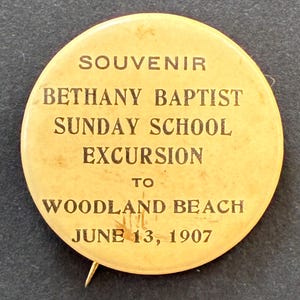May include: Vintage souvenir pin with text: "SOUVENIR BETHANY BAPTIST SUNDAY SCHOOL EXCURSION TO WOODLAND BEACH JUNE 13, 1907." The pin is round, with a cream-colored background and dark brown text. A metal pin is attached to the back.