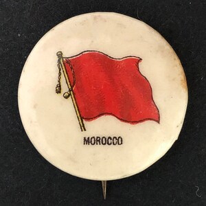 May include: A circular white pinback button featuring a graphic illustration of a red flag on a gold pole with a small chain detail. The word MOROCCO is printed in black capital letters below the flag illustration on the white background.