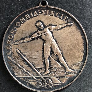 May include: A circular silver-toned metal medallion featuring a relief of a person throwing a javelin. The text LABOR OMNIA VINCIT is embossed along the top edge, and the letters B.I.K. are inscribed on a banner at the bottom with a spear.