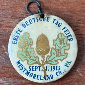 May include: A vintage round pin with the text "ERSTE DEUTSCHE TAG FEIER" in blue. The pin features a gold acorn and green oak leaves. The text "WESTMORELAND CO., PA. SEPT. 1, 1913" is also visible. A black loop is at the top.