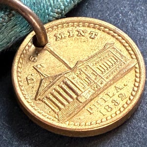May include: A gold-coloured coin with the word "MINT" at the top and "PHILA 1882" at the bottom. The coin features a building design and is attached to a teal ribbon.