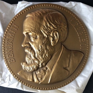 May include: A bronze-colored circular relief sculpture featuring a detailed profile of a man with a beard and suit. The name "BENJAMIN HARRISON" is inscribed around the edge. The sculpture has a textured surface and is likely a commemorative item.