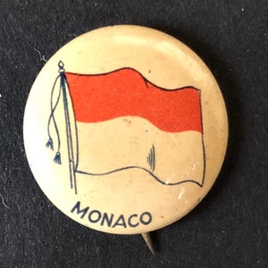 May include: A round button with a white background and a red and white flag of Monaco. The flag is waving and the button has the word "MONACO" printed in black letters below the flag.