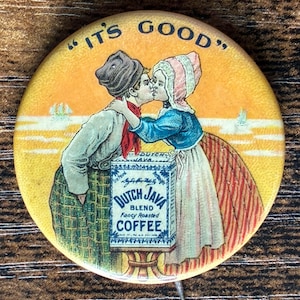 Może przedstawiać: Okrągła, zabytkowa przypinka z napisem "IT'S GOOD" u góry. Przypinka przedstawia parę w tradycyjnych holenderskich strojach całujących się, z pudełkiem "Dutch Java Blend Fancy Roasted Coffee" pomiędzy nimi. Tło jest żółte.