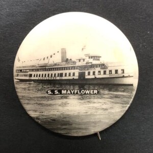 Peut inclure: Une photo en noir et blanc d'un ferry à passagers vintage nommé "S.S. Mayflower" sur l'eau. Le ferry a une coque blanche et un pont supérieur noir avec des fenêtres. La photo est sur un bouton rond.