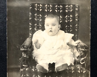 C. Foto de 1910, bebé RPPC con relicario y vestido blanco en una mecedora