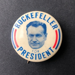 May include: A white button with a blue and red border. The button features a black and white image of a man with the text "Rockefeller for President" in blue.
