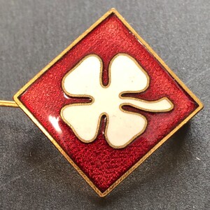 May include: A gold-colored pin with a red enamel square featuring a white four-leaf clover.