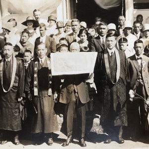 May include: A black and white photo of a large group of people, mostly adults, standing in front of a building. Many of the people are wearing traditional clothing. A person in the center of the group is holding a large, white banner.