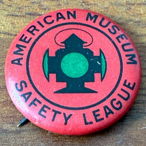 May include: A red circular pin with the text "AMERICAN MUSEUM SAFETY LEAGUE" in white lettering around the edge. The center features a black and green graphic design. A pin is attached to the back.