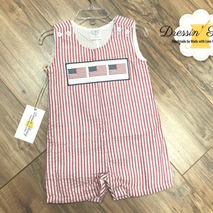 May include: Red and white striped baby overalls with a patch of three American flags on the front. The overalls are hanging on a hanger. The text "Dressin' Fancy" is visible in the top right corner of the image.
