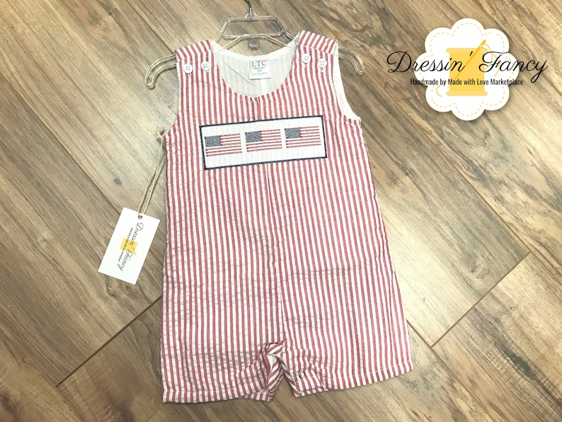 May include: Red and white striped baby overalls with a patch of three American flags on the front. The overalls are hanging on a hanger. The text "Dressin' Fancy" is visible in the top right corner of the image.
