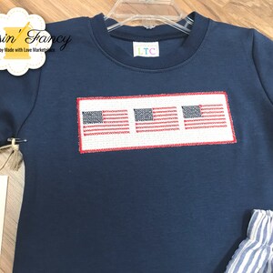 Pu&ograve; includere: Una maglietta blu navy a maniche corte con tre bandiere americane ricamate in fila. La maglietta ha un'etichetta bianca con il testo "LTC" su di essa.