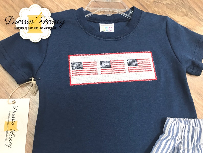 Pu&ograve; includere: Una maglietta blu navy a maniche corte con tre bandiere americane ricamate in fila. La maglietta ha un'etichetta bianca con il testo "LTC" su di essa.