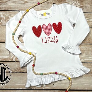 Puede incluir: Camiseta blanca de manga larga con dobladillo y pu&ntilde;os con volantes. La camiseta presenta tres corazones rojos y rosas con un nombre bordado en rojo "Lizzy" debajo. La camiseta tiene un logotipo en blanco y negro que dice "Made with Love Marketplace" en el lado izquierdo.