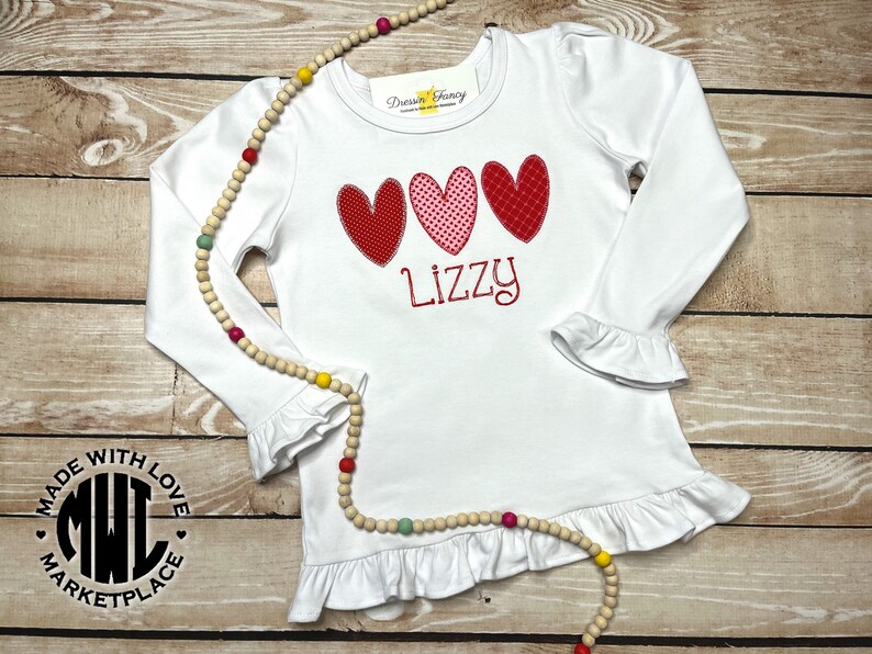 Puede incluir: Camiseta blanca de manga larga con dobladillo y pu&ntilde;os con volantes. La camiseta presenta tres corazones rojos y rosas con un nombre bordado en rojo "Lizzy" debajo. La camiseta tiene un logotipo en blanco y negro que dice "Made with Love Marketplace" en el lado izquierdo.
