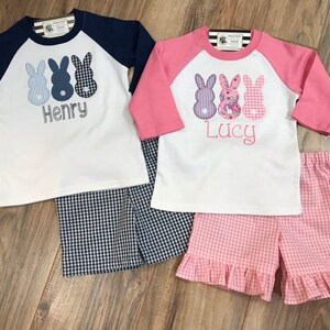 May include: Two children's outfits featuring a baseball tee shirt and gingham shorts. The shirts have a design of three Easter bunnies with the names "Henry" and "Lucy" embroidered on the front.