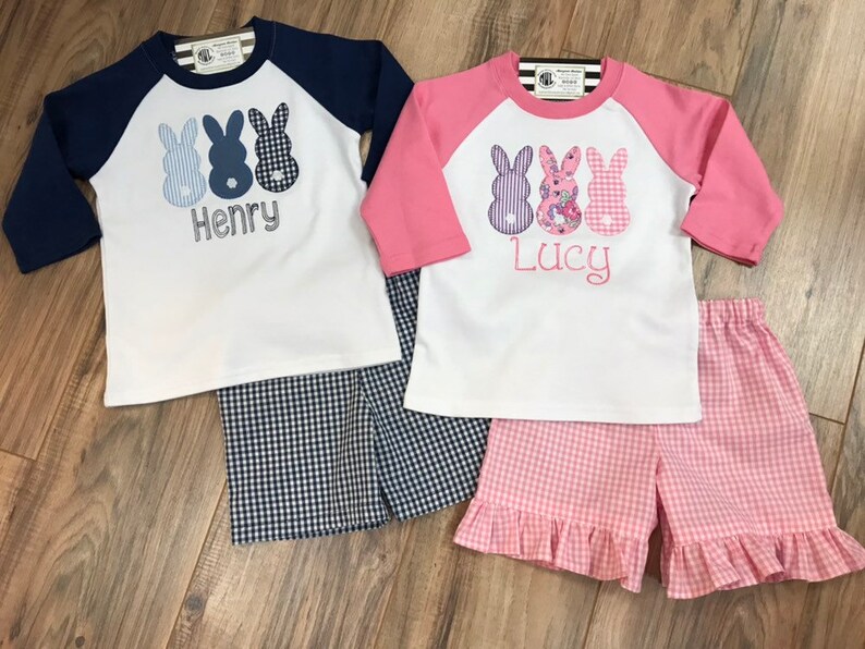 May include: Two children's outfits featuring a baseball tee shirt and gingham shorts. The shirts have a design of three Easter bunnies with the names "Henry" and "Lucy" embroidered on the front.