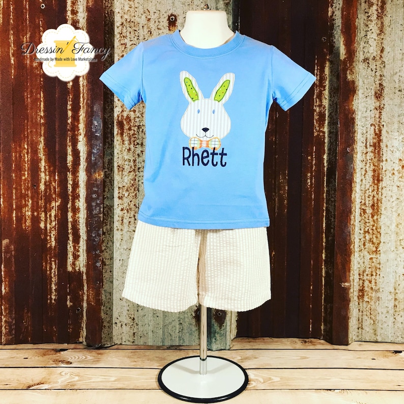 May include: A light blue t-shirt with a white bunny rabbit wearing a bow tie and the name "Rhett" embroidered on the front. The shirt is paired with beige shorts.