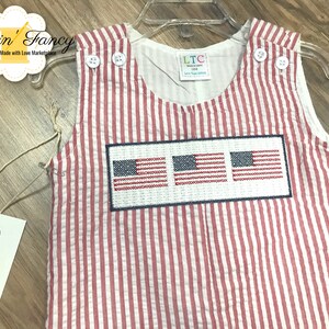 May include: Red and white striped baby overalls with three embroidered American flags on the front. The overalls have a white label with the text "LTC Little Threads Collection".