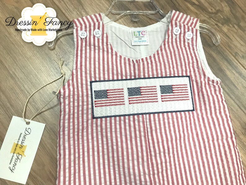 May include: Red and white striped baby overalls with three embroidered American flags on the front. The overalls have a white label with the text "LTC Little Threads Collection".