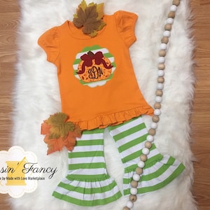 May include: A toddler's orange outfit featuring a pumpkin design and green and white striped trousers. The top has a frilled hem and short sleeves. The outfit is decorated with autumn leaves. The brand name "Dressin' Fancy" is visible.