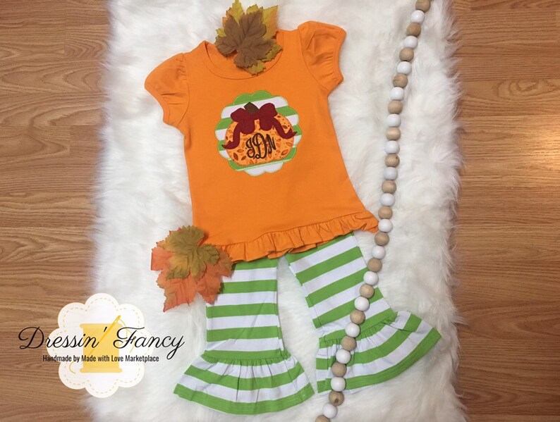 May include: A toddler's orange outfit featuring a pumpkin design and green and white striped trousers. The top has a frilled hem and short sleeves. The outfit is decorated with autumn leaves. The brand name "Dressin' Fancy" is visible.