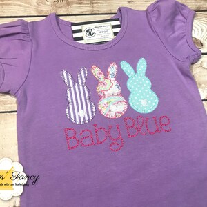 Puede incluir: Camiseta morada de manga corta con tres formas de conejo bordadas y el texto "Baby Blue".