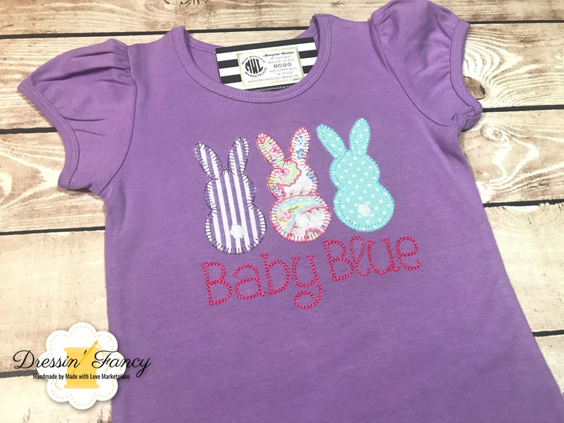 Puede incluir: Camiseta morada de manga corta con tres formas de conejo bordadas y el texto "Baby Blue".