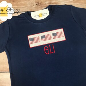 Pu&ograve; includere: Una maglietta blu navy con tre bandiere americane ricamate e il nome "Eli" in rosso.