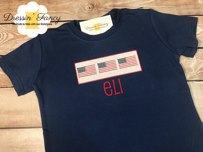 Pu&ograve; includere: Una maglietta blu navy con tre bandiere americane ricamate e il nome "Eli" in rosso.