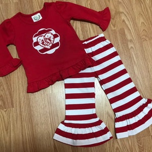K&ouml;nnte beinhalten: Rote und wei&szlig;e gestreifte Hose mit R&uuml;schen am Saum und ein rotes Langarmshirt mit einem wei&szlig;en Herz und Monogramm auf der Vorderseite.