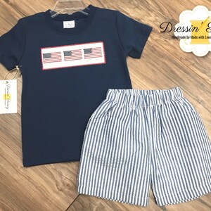 Pu&ograve; includere: Una maglietta blu navy a maniche corte con un designo ricamato bianco di tre bandiere americane. La maglietta &egrave; abbinata a pantaloncini a righe blu e bianche.