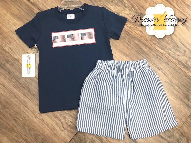 Pu&ograve; includere: Una maglietta blu navy a maniche corte con un designo ricamato bianco di tre bandiere americane. La maglietta &egrave; abbinata a pantaloncini a righe blu e bianche.