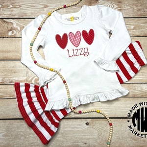 Puede incluir: Camiseta blanca de manga larga con corazones rojos, rosas y blancos y el nombre "Lizzy" bordado en la parte delantera. La camiseta tiene un dobladillo con volantes y se combina con pantalones de rayas rojas y blancas. Los pantalones tienen un dobladillo con volantes y est&aacute;n hechos de un tejido el&aacute;stico. El conjunto est&aacute; hecho con amor y es perfecto para una ni&ntilde;a peque&ntilde;a.