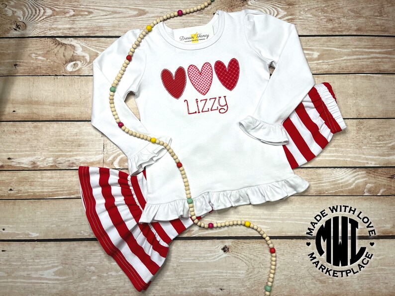 Puede incluir: Camiseta blanca de manga larga con corazones rojos, rosas y blancos y el nombre "Lizzy" bordado en la parte delantera. La camiseta tiene un dobladillo con volantes y se combina con pantalones de rayas rojas y blancas. Los pantalones tienen un dobladillo con volantes y est&aacute;n hechos de un tejido el&aacute;stico. El conjunto est&aacute; hecho con amor y es perfecto para una ni&ntilde;a peque&ntilde;a.