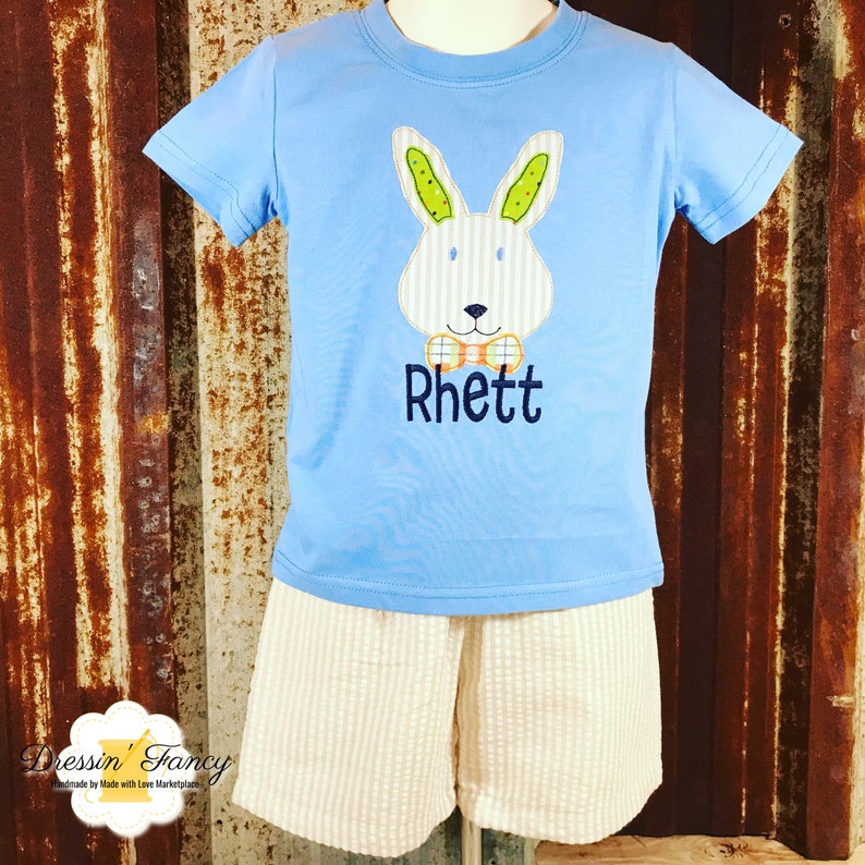 May include: A light blue t-shirt with a white bunny applique wearing a bow tie. The bunny has green ears and a striped pattern. The shirt has the name "Rhett" embroidered on the front.
