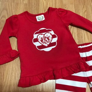 K&ouml;nnte beinhalten: Rot-wei&szlig; gestreifte Hose mit einem roten Langarm-Shirt mit einem wei&szlig;en Herz mit roten Streifen und einem Monogramm in der Mitte. Das Shirt hat R&uuml;schen an den &Auml;rmeln und am Saum.