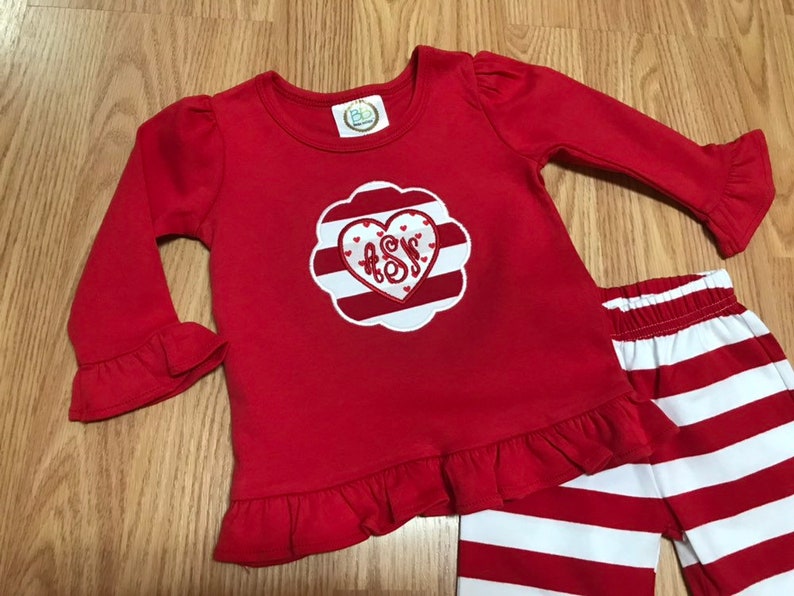 K&ouml;nnte beinhalten: Rot-wei&szlig; gestreifte Hose mit einem roten Langarm-Shirt mit einem wei&szlig;en Herz mit roten Streifen und einem Monogramm in der Mitte. Das Shirt hat R&uuml;schen an den &Auml;rmeln und am Saum.