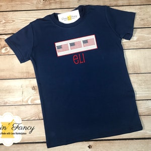 Pu&ograve; includere: Maglietta blu navy con un rettangolo bianco con tre piccole bandiere americane e il nome "ELI" ricamato in rosso.