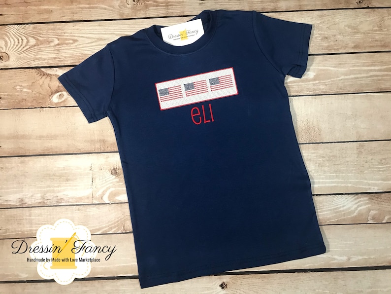 Pu&ograve; includere: Maglietta blu navy con un rettangolo bianco con tre piccole bandiere americane e il nome "ELI" ricamato in rosso.